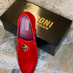 May include: A red velvet loafer with a gold-colored panther head emblem. The shoe is displayed on a stone surface next to a black box with gold lettering that reads "TERRONI ITALIAN DESIGN".