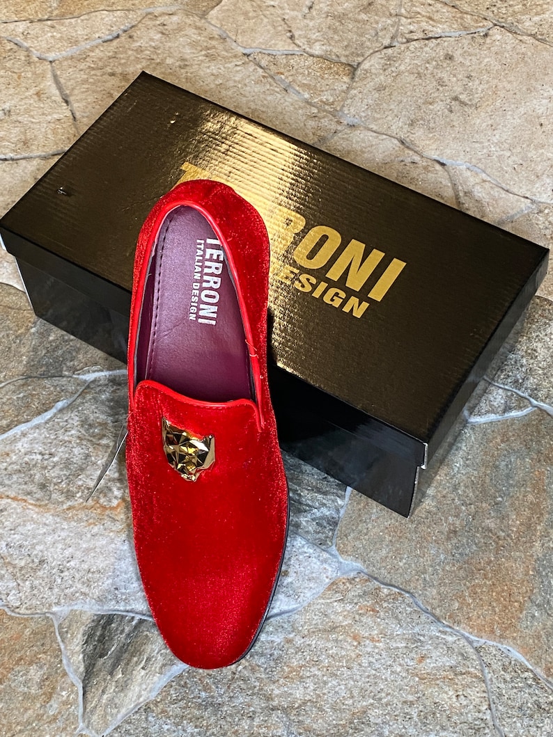 May include: A red velvet loafer with a gold-colored panther head emblem. The shoe is displayed on a stone surface next to a black box with gold lettering that reads "TERRONI ITALIAN DESIGN".