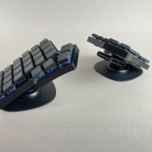Puede incluir: Un teclado mecánico ergonómico dividido que consta de dos paneles negros separados con teclas oscuras e interruptores azules visibles. Cada panel está montado sobre una base circular negra con un mecanismo de inclinación ajustable.