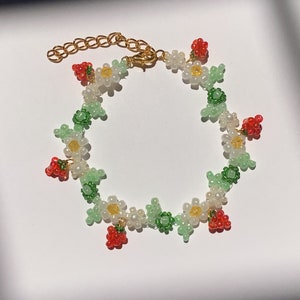 Peut inclure: Un bracelet délicat en perles avec un motif floral composé de marguerites blanches, de feuilles vertes et de fraises rouges. Le bracelet est fait de petites perles et a un fermoir doré.
