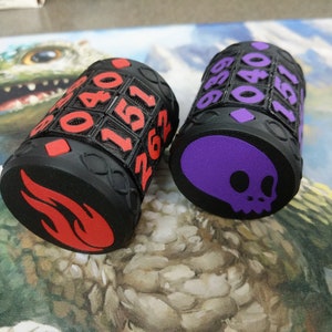May include: Two black dice with different colored symbols. One die has a red flame symbol and the other has a purple skull symbol. Both dice have numbers on the sides.