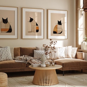 Pu&ograve; includere: Tre illustrazioni incorniciate di gatti bianco e nero con motivi geometrici in tonalit&agrave; arancio e marrone, appese a una parete beige sopra un divano angolare marrone con cuscini a righe e un tavolino da caff&egrave; in legno.