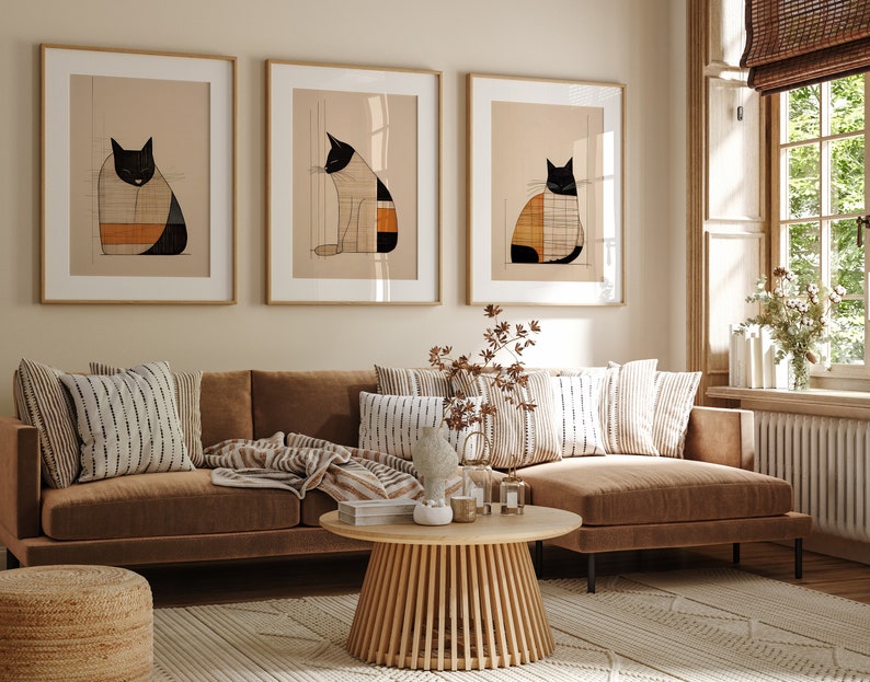 Pu&ograve; includere: Tre illustrazioni incorniciate di gatti bianco e nero con motivi geometrici in tonalit&agrave; arancio e marrone, appese a una parete beige sopra un divano angolare marrone con cuscini a righe e un tavolino da caff&egrave; in legno.