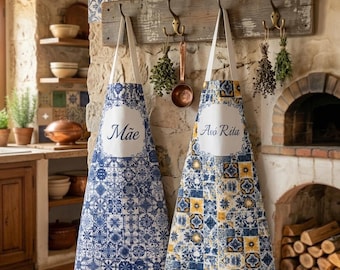 May include: Two aprons with white neck straps hang on a wooden rack. One apron is blue and white with the word "Mãe" in a white oval. The other is yellow, blue, and white with "Avó Rita" in a white oval. The aprons have a tile-like pattern.