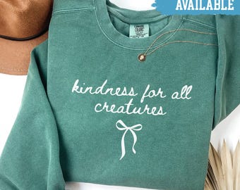 Könnte beinhalten: Ein hellgrünes Sweatshirt mit dem weißen Schriftzug "kindness for all creatures". Unter dem Text befindet sich eine weiße Schleifengrafik. Die Worte "Shirts Available" stehen in einem blauen Banner oben. Eine goldene Halskette ist sichtbar.