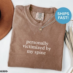 Peut inclure: T-shirt couleur expresso avec l'inscription blanche « personally victimized by my spine ». Le t-shirt a un col rond et des manches courtes. Un collier argenté avec un petit pendentif est sur le t-shirt.