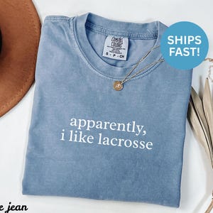 May include: A blue jean colored t-shirt with the text "apparently, i like lacrosse" in white. The shirt has a crew neck and short sleeves. A gold necklace with a pendant is on the shirt.