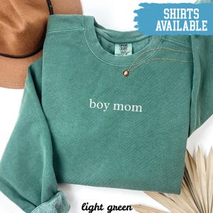 May include: Light green sweatshirt with the text "boy mom" in white. A gold necklace with a pendant is draped on the shirt. The words "SHIRTS AVAILABLE" are in a blue banner in the upper right corner.