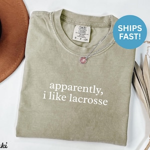 May include: Khaki-colored t-shirt with the text "apparently, i like lacrosse" in white. The shirt has a crew neck and a small tag on the upper left. A silver necklace with a pendant is visible.