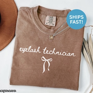May include: Espresso-colored t-shirt with the words "eyelash technician" in white script and a bow graphic. The shirt has a crew neck and a Comfort Colors label. A silver necklace with a pendant is visible.