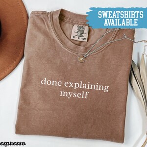 May include: Espresso-colored t-shirt with the phrase "done explaining myself" in white. The shirt has a crew neck and short sleeves. A silver necklace with a pendant is draped across the shirt. The words "SWEATSHIRTS AVAILABLE" are in a blue banner.
