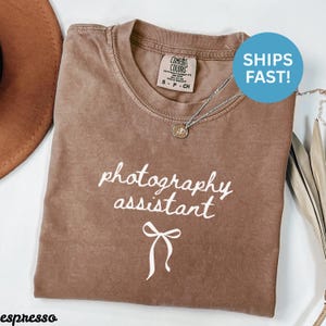 May include: Espresso-colored t-shirt with the words "photography assistant" in white script. A silver necklace with a small pendant is draped on the shirt. The shirt has a crew neck and short sleeves.