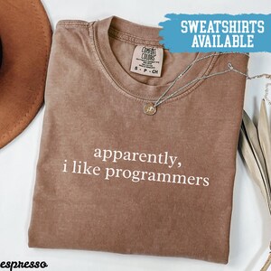 May include: Espresso-colored t-shirt with the text "apparently, i like programmers" in white. The shirt has a crew neck and is made of a soft material. A silver necklace with a small pendant is draped across the shirt.
