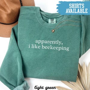 May include: Light green sweatshirt with the text "apparently, i like beekeeping" in white. The shirt has a crew neck and long sleeves. A gold necklace with a pendant is visible. The words "Shirts Available" are in a blue banner.