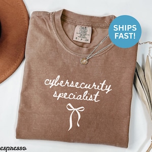 Könnte beinhalten: Espressofarbenes T-Shirt mit dem weißen Schriftzug "cybersecurity specialist" und einer Schleifengrafik. Das Shirt hat einen Rundhalsausschnitt und kurze Ärmel. Eine silberne Halskette mit Anhänger ist sichtbar. Das Shirt ist mit "Comfort Colors" gekennzeichnet.