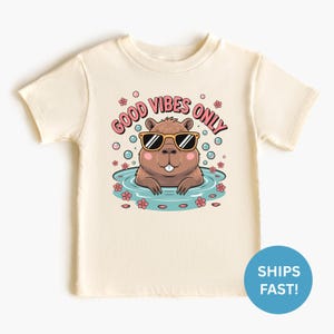 May include: Cream-colored t-shirt featuring a cartoon capybara wearing sunglasses and relaxing in a pool. The shirt has the text "GOOD VIBES ONLY" in pink and black. Pink flowers and bubbles surround the capybara.