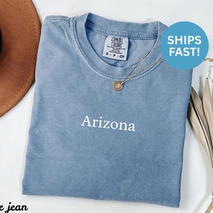 May include: A blue jean Comfort Colors t-shirt with the word "Arizona" printed in white. The shirt has a crew neck and short sleeves. A gold necklace with a pendant is draped on the shirt. A brown hat is visible in the background.