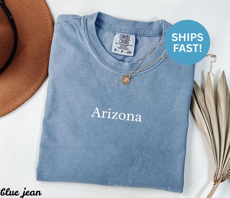 May include: A blue jean Comfort Colors t-shirt with the word "Arizona" printed in white. The shirt has a crew neck and short sleeves. A gold necklace with a pendant is draped on the shirt. A brown hat is visible in the background.