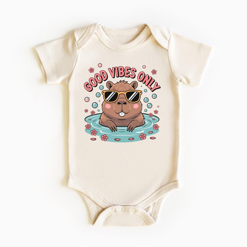 May include: Cream-colored baby bodysuit with a cartoon graphic. The graphic features a brown beaver wearing sunglasses in a pool of water, surrounded by bubbles and flowers. Text reads "GOOD VIBES ONLY" in pink.