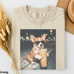 Op de afbeelding: Ivoorkleurig T-shirt met een afbeelding van een corgi met een zonnebril die ijskoffie drinkt. De corgi zit op een richel met een vage stadsachtergrond. Het shirt heeft een Comfort Colors-label.