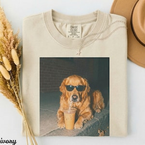 Cream t-shirt with golden retriever wearing sunglasses holding iced coffee.