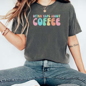 Pode incluir: T-shirt cinzenta escura com o texto "Often Yaps About Coffee" em letras coloridas de estilo retro. A camisa tem um corte folgado e parece ser feita de material macio. A estética geral é casual e confortável.