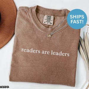 Könnte beinhalten: Espressofarbenes Comfort Colors T-Shirt mit dem weißen Text "readers are leaders". Das Shirt hat einen Rundhalsausschnitt und kurze Ärmel. Eine silberne Halskette mit einem kleinen Anhänger befindet sich auf dem Shirt. Ein brauner Hut ist in der oberen linken Ecke.