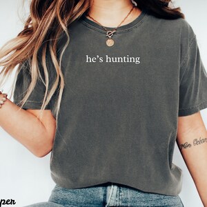 May include: Dark gray t-shirt with the text "he's hunting" in white. The shirt has short sleeves and a crew neck. The model is wearing a gold necklace and a bracelet.