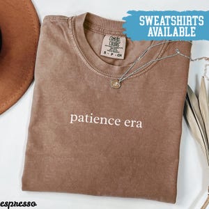 May include: Espresso-colored t-shirt with the words "patience era" in white. A silver necklace with a small pendant rests on the shirt. The shirt is labeled "Comfort Colors" and the text "SWEATSHIRTS AVAILABLE" is in a blue banner.