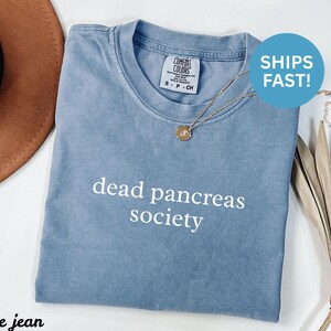 Puede incluir: Una camiseta azul con la frase "dead pancreas society" en blanco. La camiseta tiene cuello redondo y mangas cortas. Un collar dorado con un colgante est&aacute; sobre la camiseta. Un c&iacute;rculo azul con las palabras "SHIPS FAST!" est&aacute; en la camiseta.