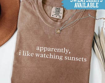 May include: Espresso-colored t-shirt with the text "apparently, i like watching sunsets" in white. A silver necklace with a pendant rests on the shirt. The shirt is labeled "Comfort Colors" and the text "SWEATSHIRTS AVAILABLE" is in a blue banner.