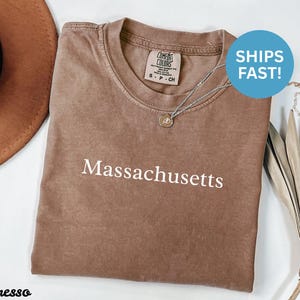 May include: Espresso-coloured t-shirt with the word "Massachusetts" printed in white. The shirt has a crew neck and a Comfort Colors tag. A silver necklace with a small pendant is draped on the shirt.