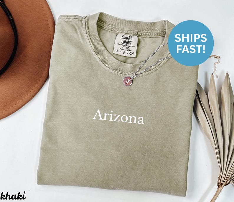 May include: Khaki-colored Comfort Colors t-shirt with the word "Arizona" printed in white. A silver necklace with a pendant is visible. A brown hat is partially visible in the upper left corner.