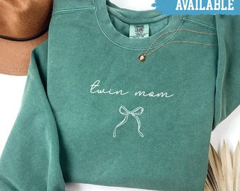 May include: Light green sweatshirt with the text "twin mom" and a bow graphic. The shirt has a crew neck and long sleeves. A gold necklace with a pendant is visible. The words "SHIRTS AVAILABLE" are in a blue banner.