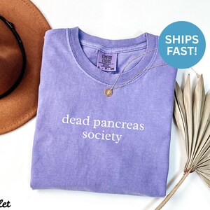 Puede incluir: Una camiseta violeta con las palabras "dead pancreas society" impresas en blanco. La camiseta tiene cuello redondo y una peque&ntilde;a etiqueta en el cuello. Un collar de oro con un colgante est&aacute; sobre la camiseta.