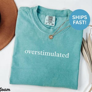 Puede incluir: Camiseta verde mar con la palabra "overstimulated" en blanco. La camiseta tiene cuello redondo y mangas cortas. Un collar dorado con un colgante est&aacute; sobre la camiseta. Un sombrero marr&oacute;n est&aacute; en el fondo.