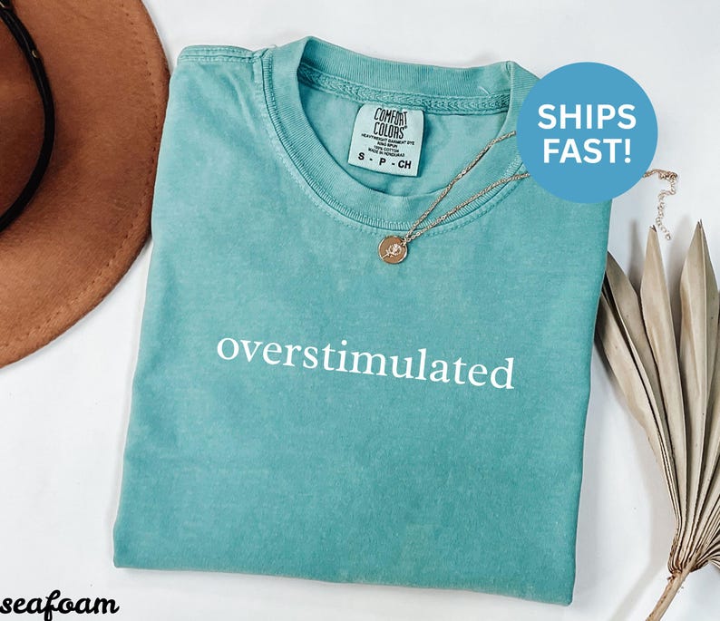 Puede incluir: Camiseta verde mar con la palabra "overstimulated" en blanco. La camiseta tiene cuello redondo y mangas cortas. Un collar dorado con un colgante est&aacute; sobre la camiseta. Un sombrero marr&oacute;n est&aacute; en el fondo.