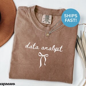 Peut inclure: T-shirt couleur expresso avec l'inscription "data analyst" en écriture blanche et un motif de nœud. Le t-shirt a un col rond et des manches courtes. Un collier argenté avec un pendentif est posé sur le t-shirt.