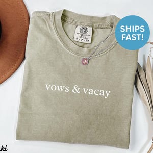 Puede incluir: Camiseta caqui con las palabras "vows & vacay" impresas en blanco. La camiseta tiene cuello redondo y mangas cortas. Se ve un collar plateado con un colgante. Un sombrero marr&oacute;n est&aacute; en el fondo.