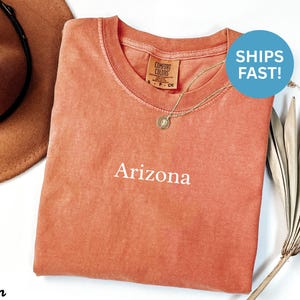 May include: A coral-colored t-shirt with the word "Arizona" in white script. The shirt has a crew neck and a small tag. A gold necklace with a pendant is on the shirt. A brown hat is in the background.