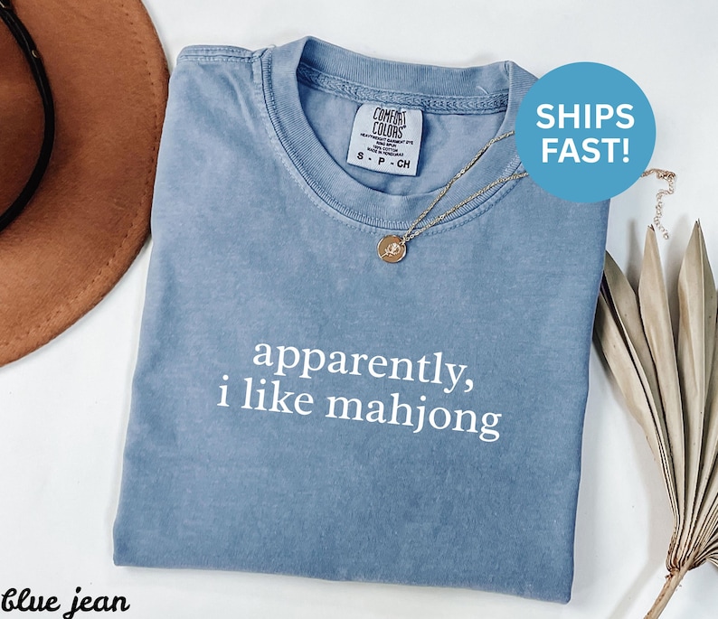 May include: A blue jean colored t-shirt with the text "apparently, i like mahjong" in white. The shirt has a crew neck and short sleeves. A gold necklace with a pendant is on the shirt.