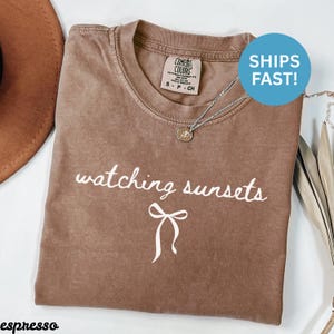 May include: Espresso-colored t-shirt with the words "watching sunsets" in white script and a bow graphic. The shirt has a crew neck and short sleeves. A silver necklace with a pendant is draped on the shirt.