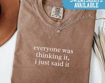 May include: Espresso-colored t-shirt with the text "everyone was thinking it, i just said it" in white. The shirt has a crew neck and a Comfort Colors tag. A silver necklace with a pendant is draped across the shirt. The words "SWEATSHIRTS AVAILABLE" are in a blue banner.