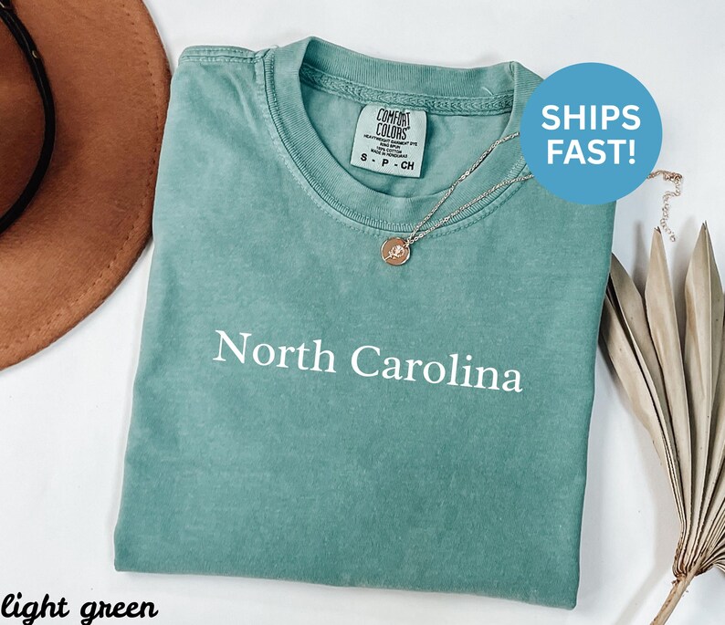 May include: Light green t-shirt with the words "North Carolina" printed in white. The shirt has a crew neck and short sleeves. A silver necklace with a small pendant is draped over the shirt. A brown hat is in the background.