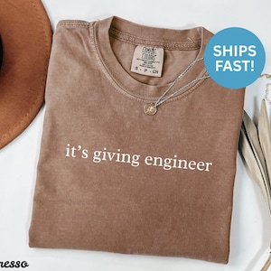 Könnte beinhalten: Espressofarbenes T-Shirt mit dem weißen Aufdruck "it's giving engineer". Das Shirt hat einen Rundhalsausschnitt und kurze Ärmel. Eine silberne Halskette mit Anhänger liegt auf dem Shirt.