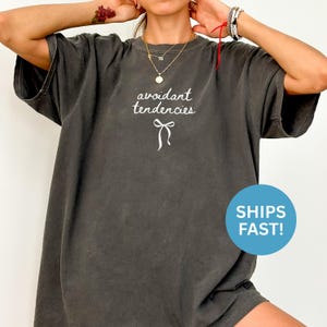 May include: Dark gray oversized t-shirt with the text "avoidant tendencies" in a white script font, and a white bow graphic. The shirt has a relaxed fit and short sleeves.
