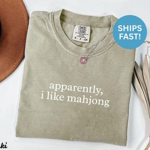 May include: Khaki-colored t-shirt with the text "apparently, i like mahjong" in white. The shirt has a crew neck and short sleeves. A silver necklace with a pink pendant is on the shirt.