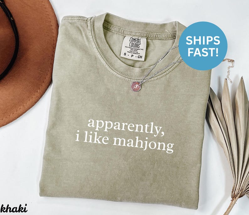 May include: Khaki-colored t-shirt with the text "apparently, i like mahjong" in white. The shirt has a crew neck and short sleeves. A silver necklace with a pink pendant is on the shirt.