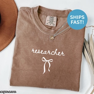 May include: Espresso-colored t-shirt with the word "researcher" in white script and a white bow graphic. The shirt has a crew neck and short sleeves. A silver necklace with a pendant is visible.