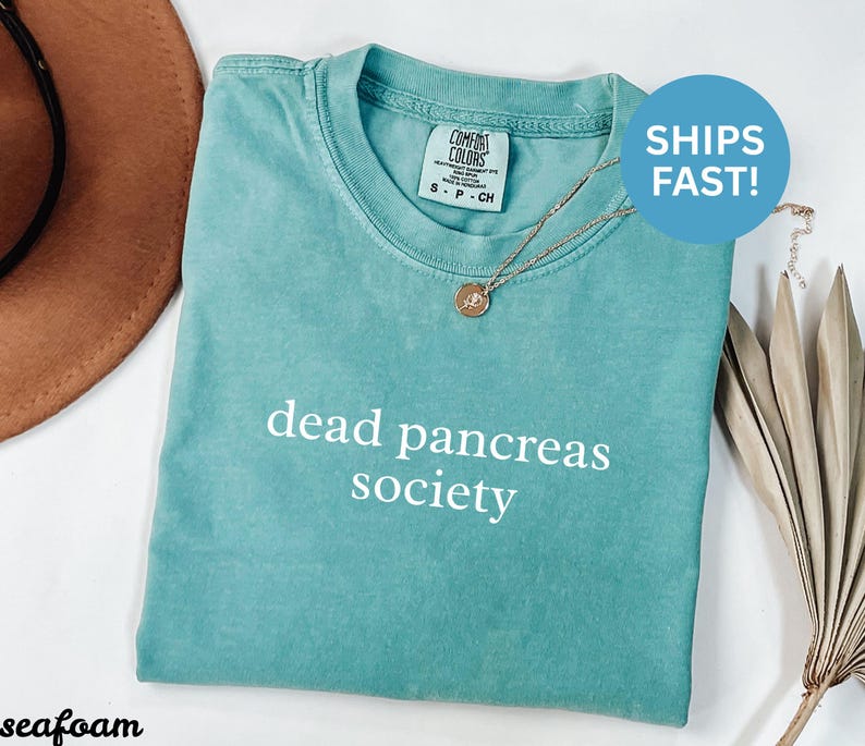 Puede incluir: Camiseta verde mar con las palabras "dead pancreas society" impresas en blanco. La camiseta tiene cuello redondo y mangas cortas. Un collar de oro con un colgante est&aacute; sobre la camiseta. Un sombrero marr&oacute;n est&aacute; en el fondo.